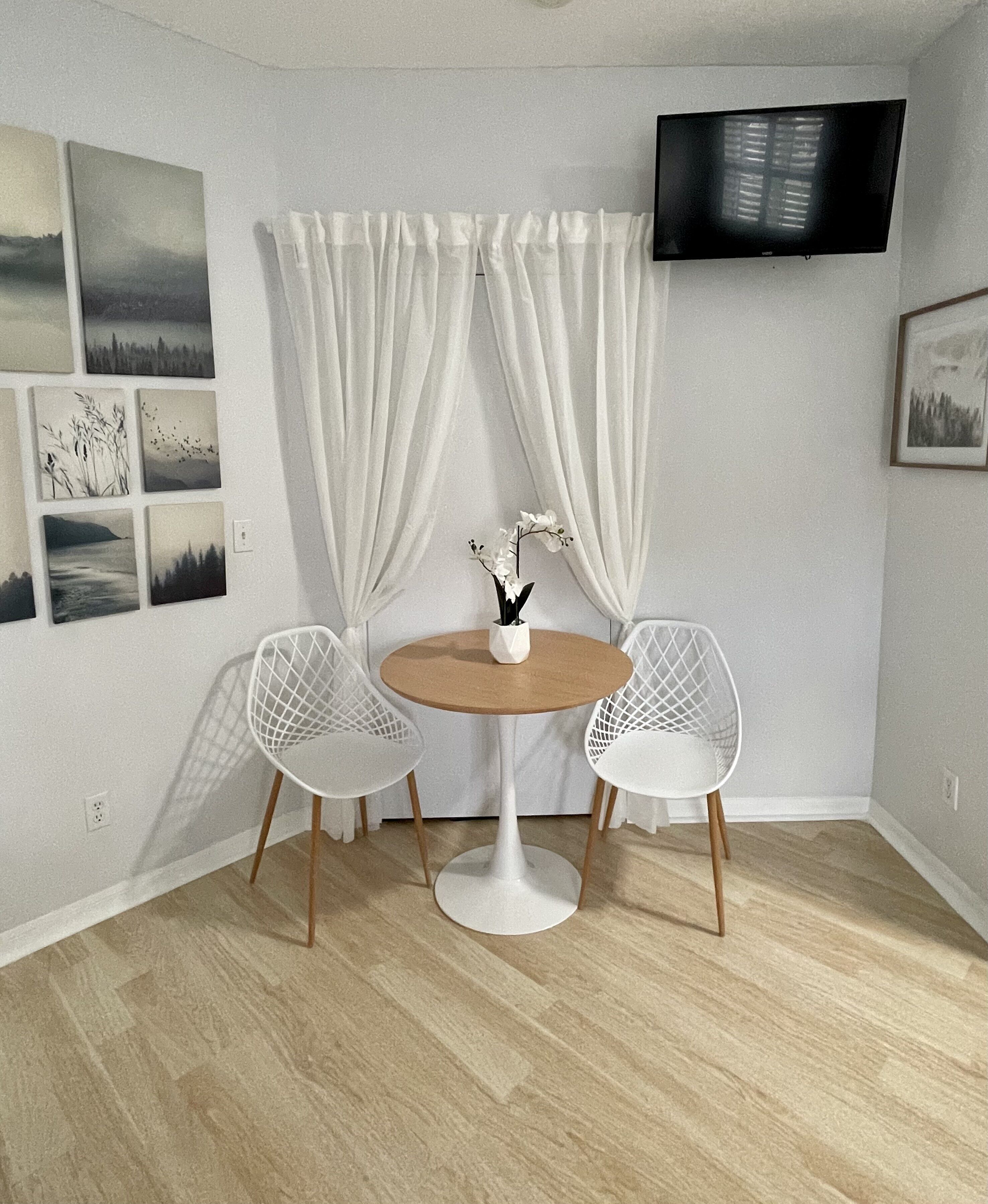 A small room with a round table, two white chairs, a flat-screen TV mounted on the wall, and several framed pictures on the wall.