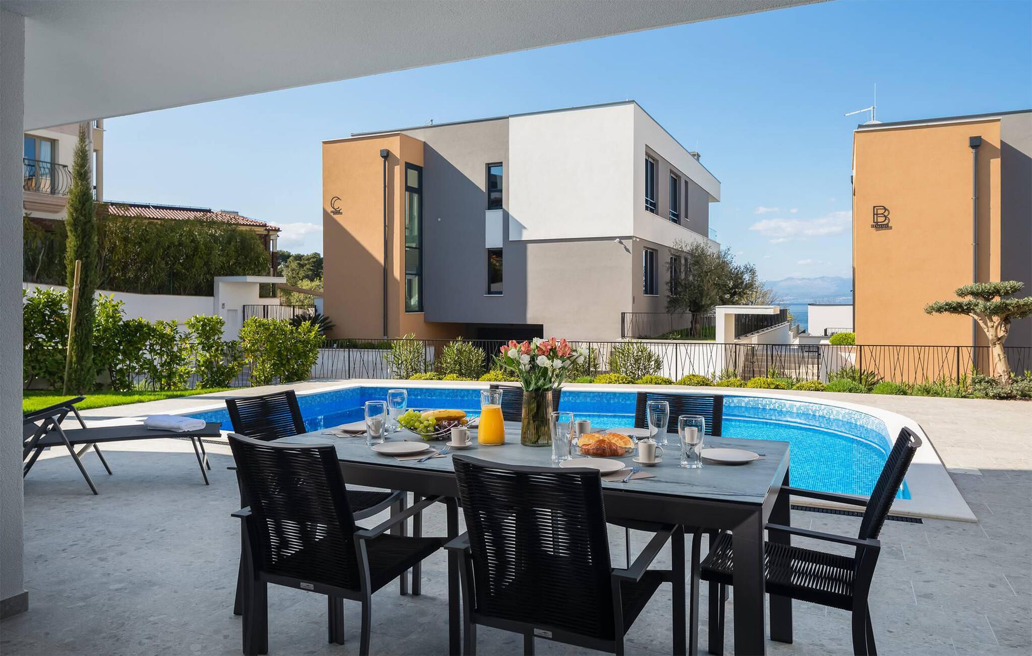 Un área de comedor junto a la piscina con una mesa preparada para cuatro personas, rodeada por una piscina y edificios modernos al fondo.