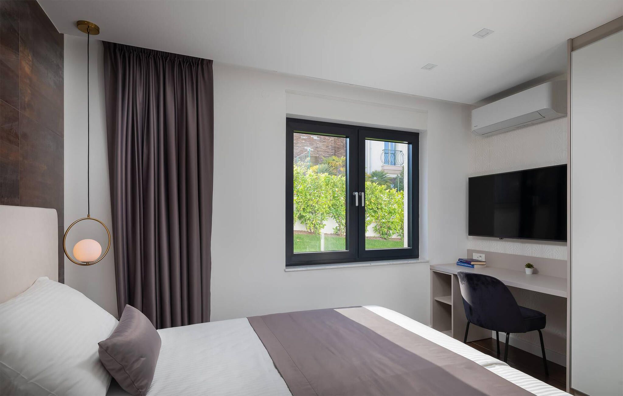 A modern hotel room with a large bed, a desk, a chair, a TV, and a window with a view of greenery.