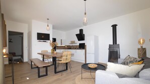 Un salon moderne comprenant un canapé, une table à manger avec des chaises, un téléviseur fixé au mur, une cheminée et un coin cuisine avec des placards et une cuisinière.