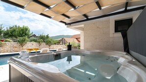 A hot tub with a stone wall backdrop, a stone patio, and a view of a pool and greenery.