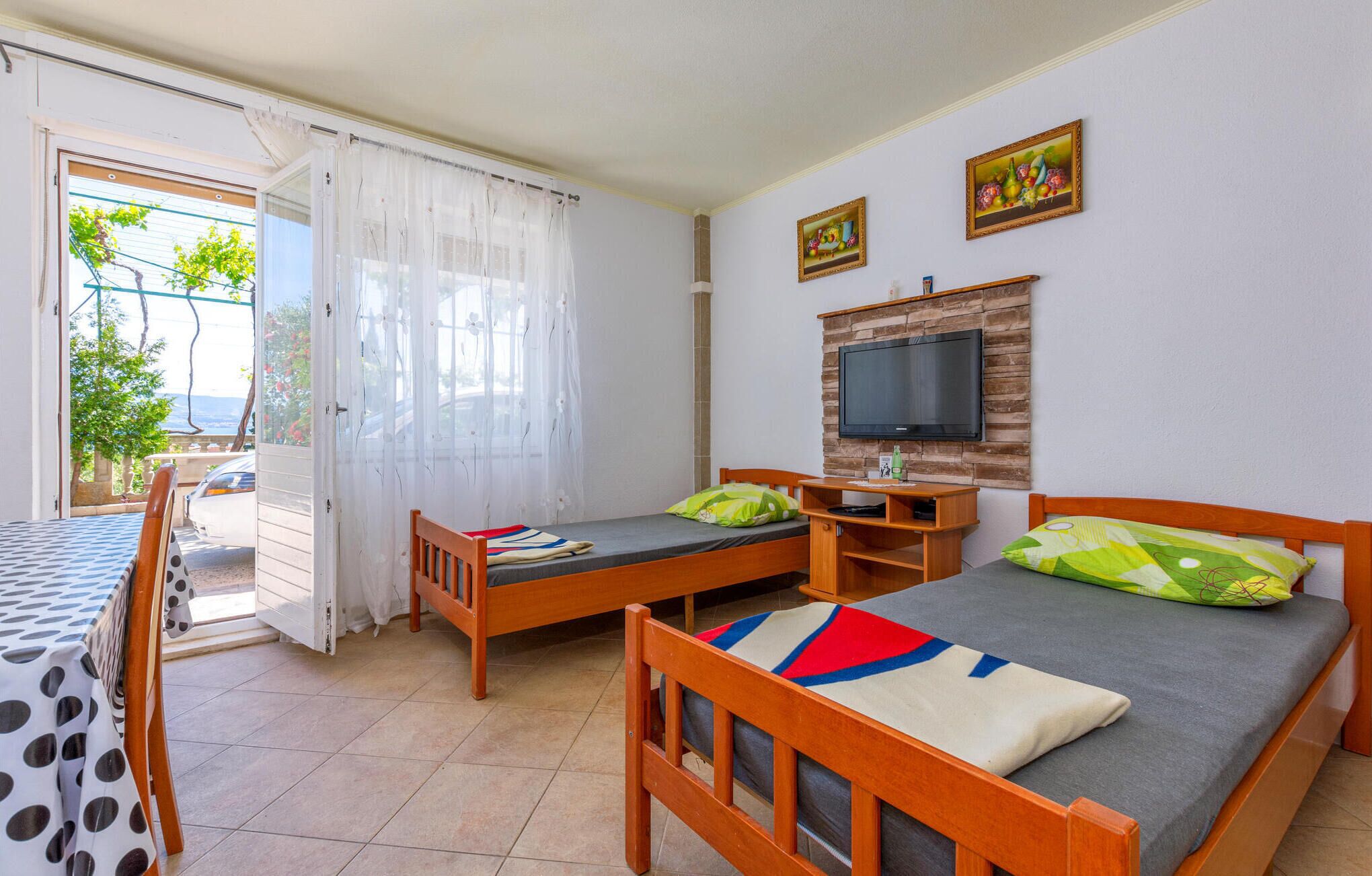 A room with two wooden beds, a TV mounted on a wooden panel, a table with a polka dot tablecloth, and a view of the outdoors through a glass door.