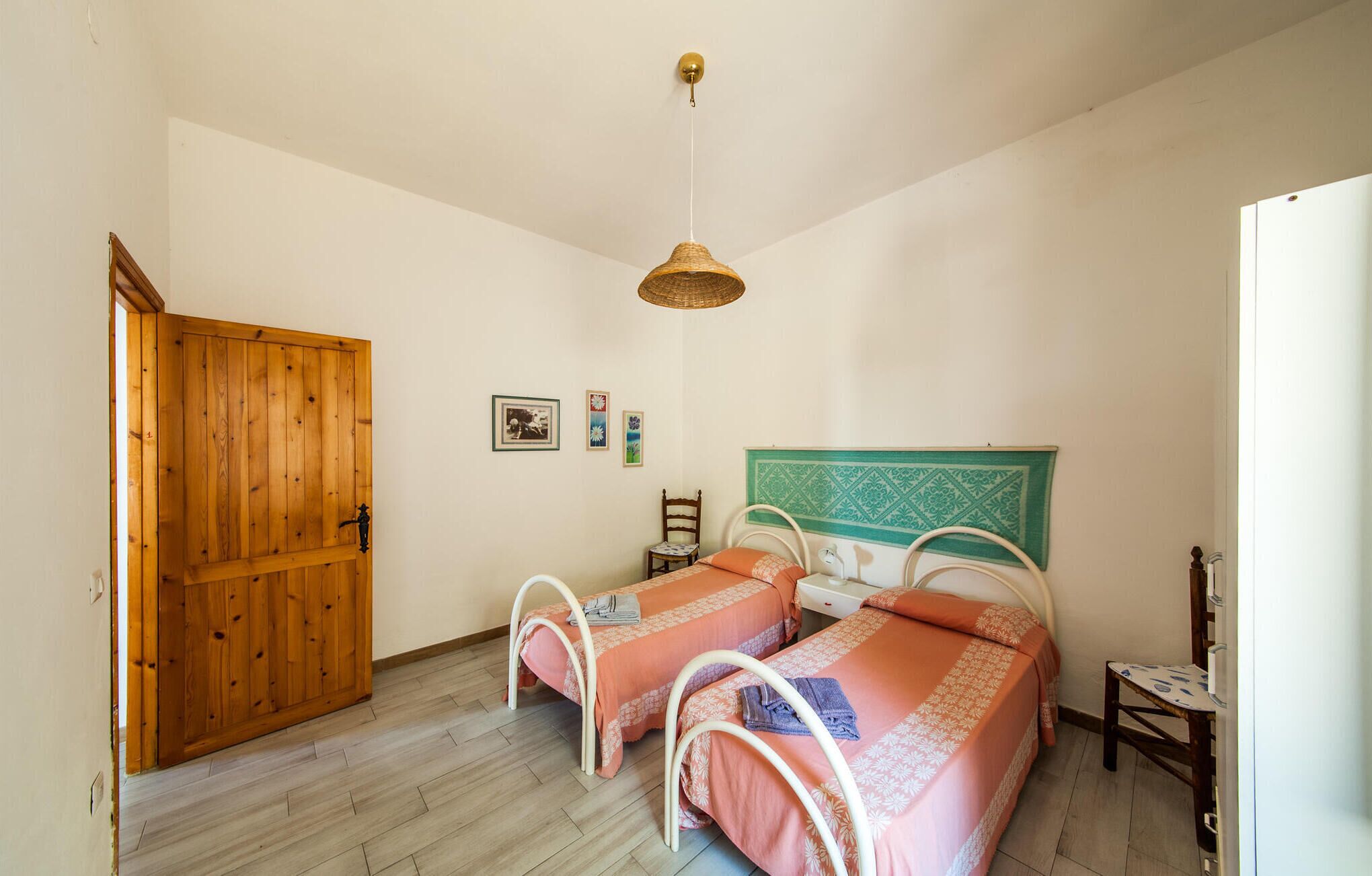 A room with two beds, a wooden door, a hanging light fixture, and a small table.