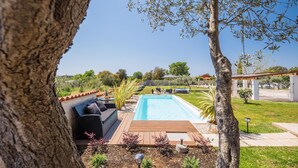 Un espace piscine doté d’une terrasse en bois, d’un coin salon avec des coussins et d’une vue sur le paysage environnant.