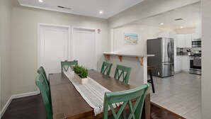 Uma área de refeições com mesa de madeira, cadeiras verdes e um arranjo de plantas. Uma cozinha visível ao fundo possui eletrodomésticos modernos.