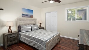 Um quarto com cama, mesas de cabeceira, ventoinha no teto, janela com persianas e uma porta.