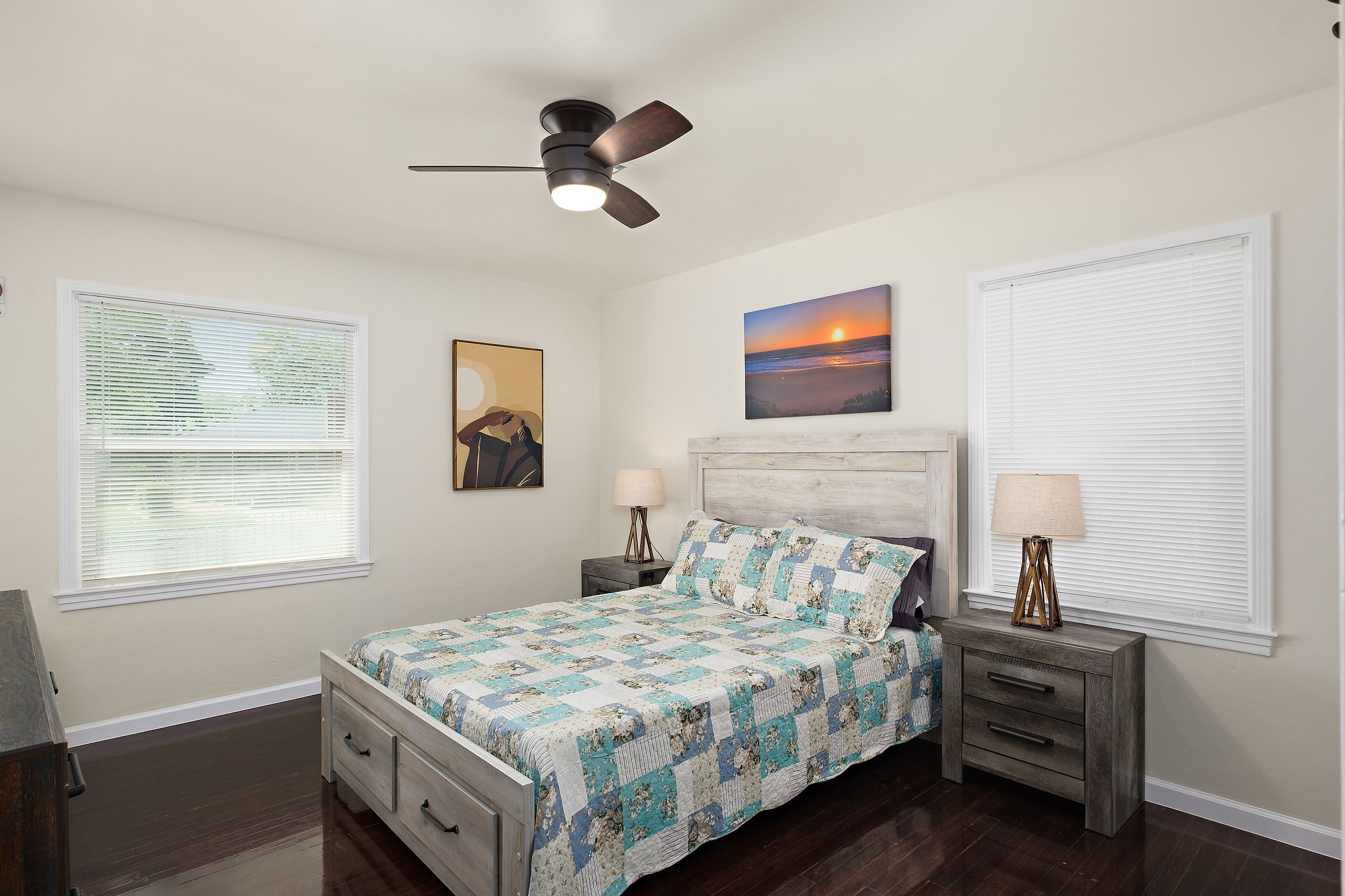 A bedroom with a bed, two bedside tables, a ceiling fan, and two windows with blinds.