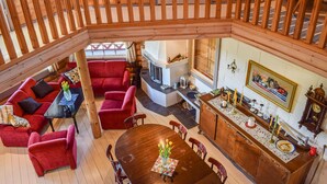 Een ruime loft-achtige kamer met een hoge plafond en zichtbare houten balken. Het heeft een gezellige zithoek met rode bekleden banken en fauteuils, een houten eettafel met een bloevas, en een keuken met een ingebouwde kookplaat en een groot raam.