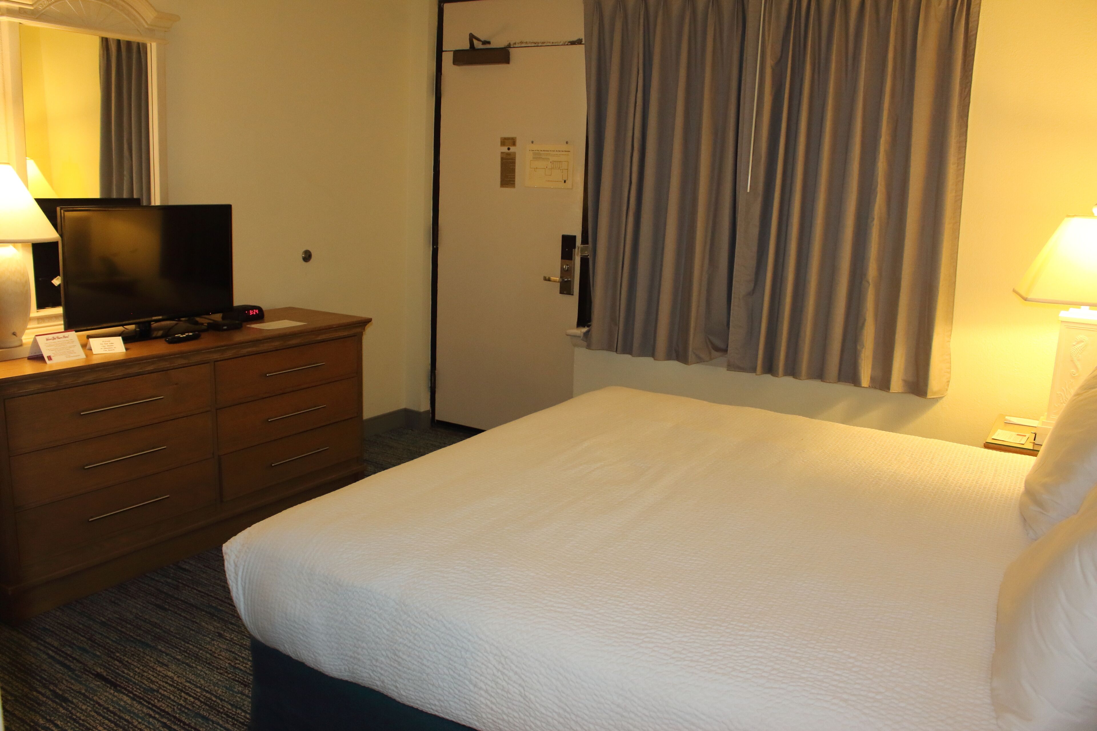 A hotel room with a bed, a TV on a wooden dresser, a desk with a lamp, and a door with a curtain.