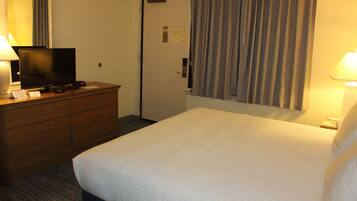 A hotel room with a bed, a TV on a wooden dresser, a desk with a lamp, and a door with a curtain.
