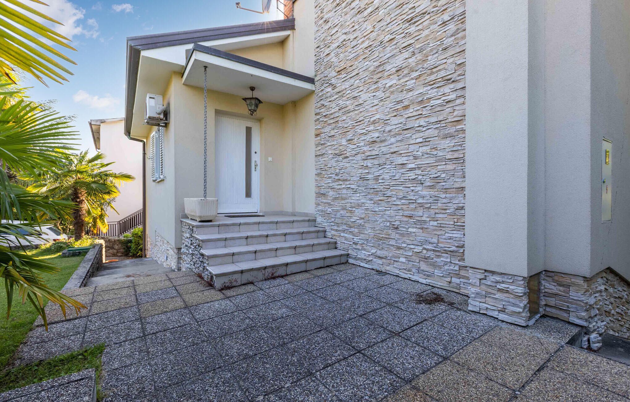 Ein modernes Gebäude mit einer Steinfassade, eine Treppe, die zu einer Tür führt, und ein kleiner Gartenbereich mit Palmen.