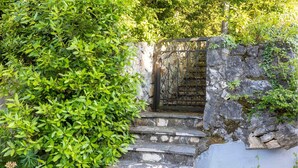 Uma escadaria de pedra leva a um portão de metal, cercada por vegetação exuberante.