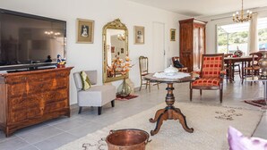 Een ruime woonkamer met een grote flatscreen televisie, een houten kast, een grijze fauteuil, een rode gestoffeerde stoel, en een eethoek met een kroonluchter.