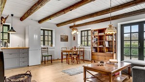 Een ruime keuken met houten balken, een eethoek met een tafel en stoelen, een boekenkast en uitzicht door grote ramen.
