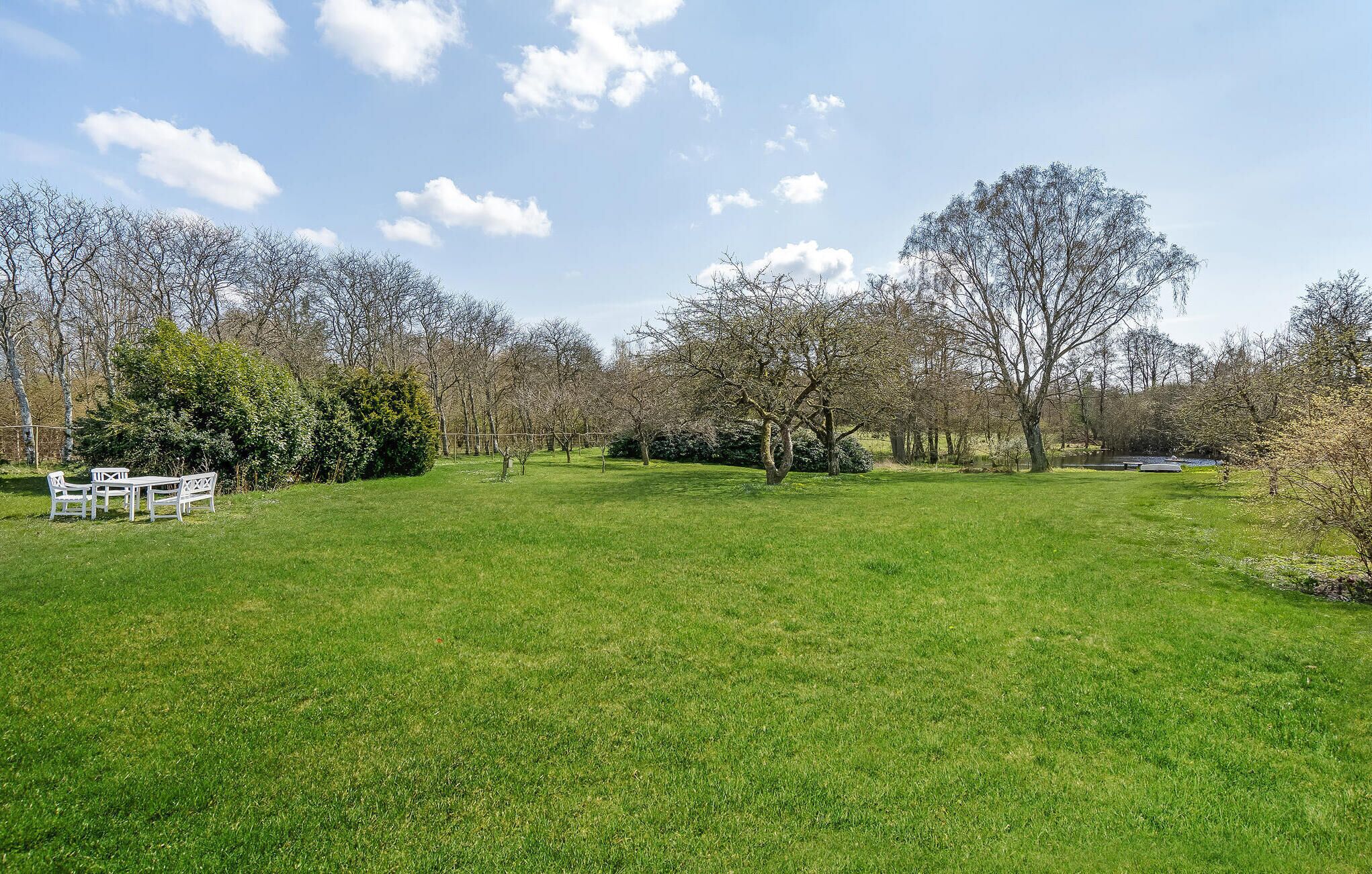 A grassy outdoor area with a white table and chairs, surrounded by trees and a fence.