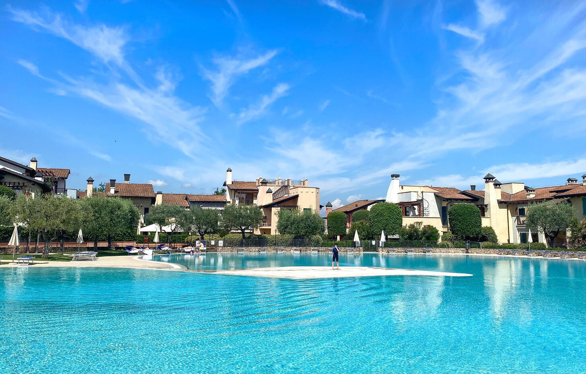 A large outdoor swimming pool with clear blue water, surrounded by buildings with Mediterranean-style architecture.