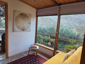 Una stanza con un'ampia finestra, un soffitto in legno, un'opera d'arte tondeggiante al muro, un piccolo tavolo con dei libri, un divano giallo e un tappeto rosso decorato.