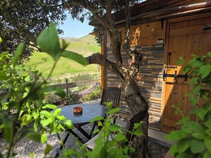 Una baita di legno dall'aspetto rustico, un tavolo rotondo con una ciotola di rame e vista su un paesaggio verde.