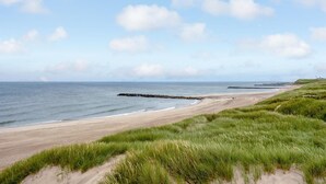 Una spiaggia con cielo sereno, dune erbose e un molo roccioso.