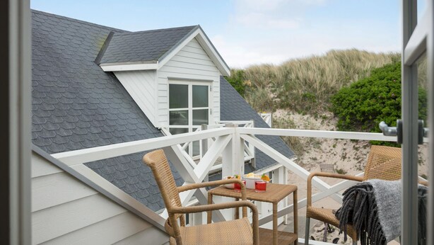 Una casa sulla spiaggia con facciata bianca, tetto grigio e un balcone con sedie in vimini e un tavolo.
