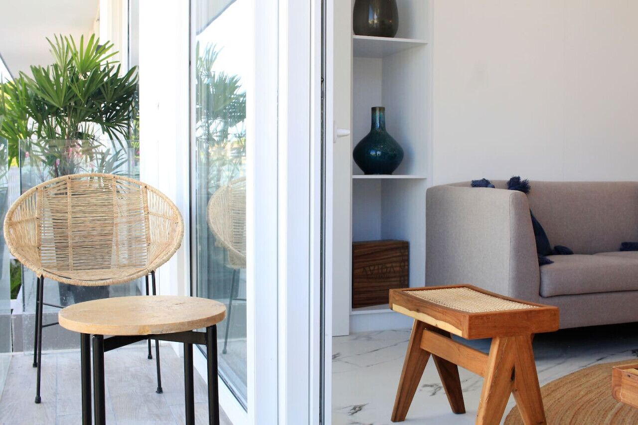 A modern living room with a wicker chair, a wooden table, a grey sofa, and a green vase on a shelf.