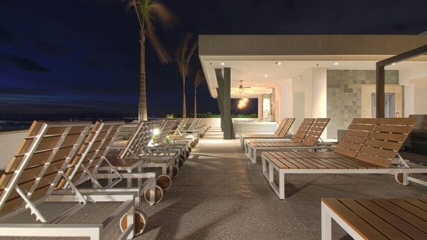 A rooftop lounge with sun loungers and a pool area, featuring a modern architectural design and a view of the ocean at night. - Avida - Stunning Rooftop Pool and Jacuzzi (Puerto Vallarta)