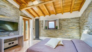 Ein Schlafzimmer mit Steinsandsteinwand, Holzbalkendecke, einem Bett, einem Fernseher und einem kleinen Fenster mit Pflanzen.