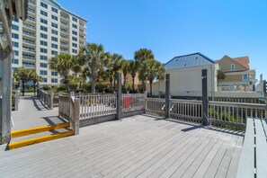 A wooden deck with a metal railing, palm trees, and a building with a blue roof in the background.