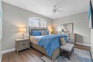 Une chambre à coucher comprenant un lit, des tables de chevet, un banc, un ventilateur de plafond, une fenêtre avec des stores et un miroir.