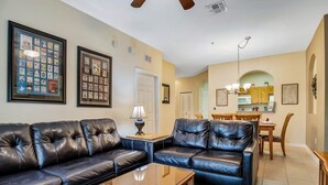 A living room with two black leather sofas, a glass coffee table, framed pictures on the walls, a dining area with a wooden table and chairs, and a kitchen visible through an archway.