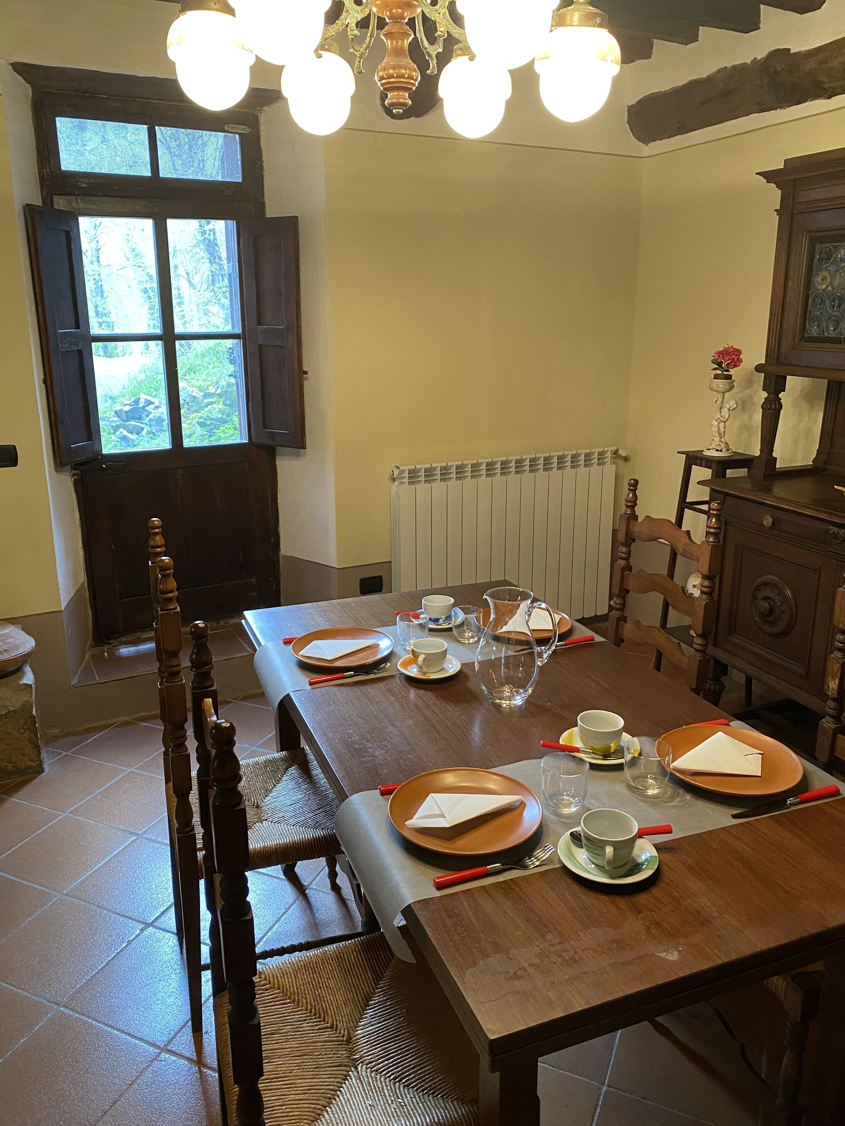 A dining room with a wooden table set for four, a radiator, a window with curtains, and a chandelier.