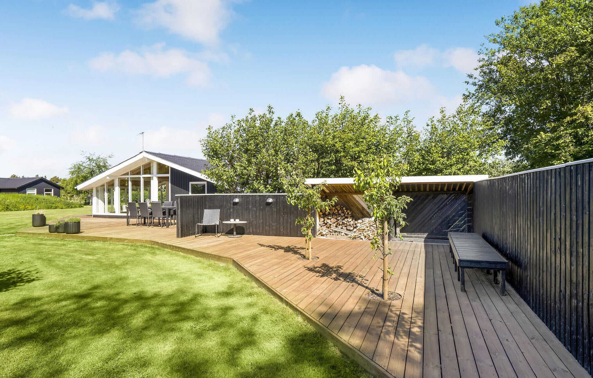 Une terrasse extérieure moderne, dotée d’un plancher en bois, entourée d’une clôture en bois et d’une pelouse verdoyante.