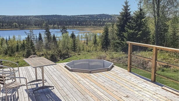 Una terraza de madera con un jacuzzi, zona de estar y vistas a un lago rodeado de árboles.