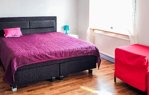 Ein Schlafzimmer mit einem Bett, einem Nachttisch, einer Lampe, einem Stuhl und einem Fenster.