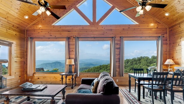 Interior de una cabaña de madera con comedor, sofá y ventanas amplias con vistas a la montaña.