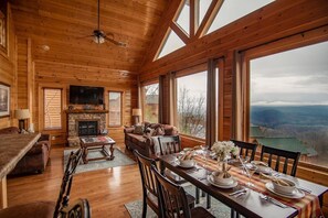 Cozy cabin with a high ceiling and exposed beams, featuring a fireplace, comfortable seating, and a dining table.