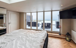 Ein Schlafzimmer mit einem großen Bett, einem Fernseher und Blick auf einen Strand durch raumhohe Fenster.