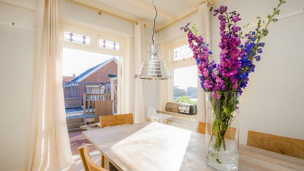 A dining area with a wooden table and chairs, a vase of flowers, and a view of a building outside. - Stunning home in Bergen aan Zee (Bergen aan Zee)