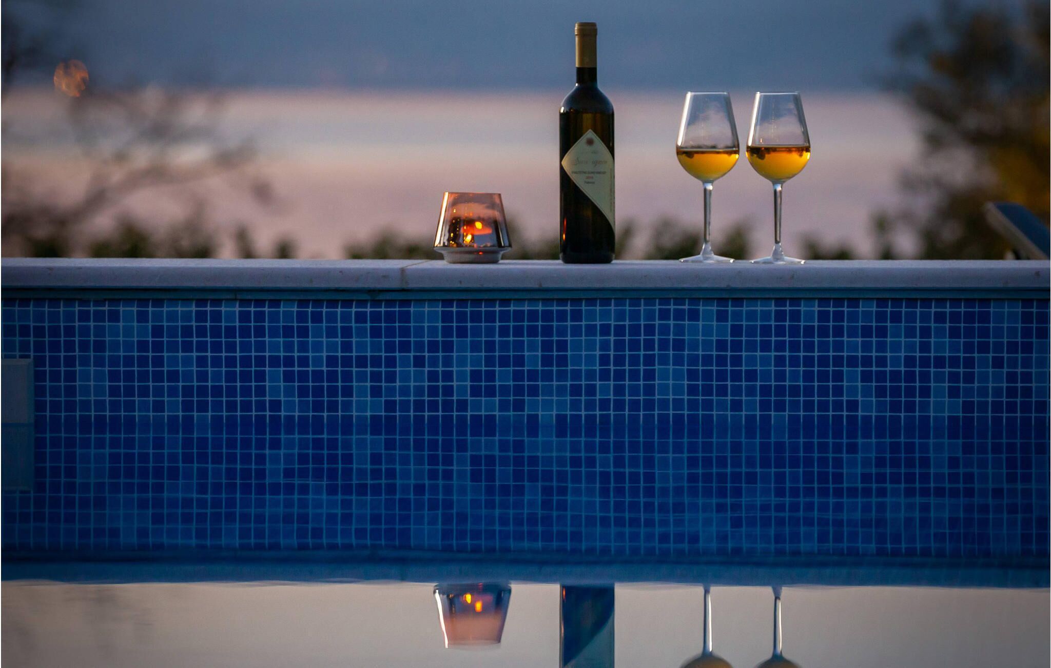A poolside setting with a bottle and two glasses of wine, reflecting on the pool's surface.