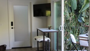 Ein kleines Zimmer mit einer weißen Tür, einem an der Wand montierten Flachbildfernseher, einem kleinen Tisch mit einer Topfpflanze, zwei Hocker mit weißen Fellüberwürfen und einem Stuhl mit weißem Kissen.