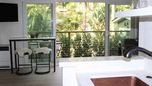 Eine moderne Küche mit einem großen Fenster mit Blick auf einen üppigen Garten. Zwei Barhocker und ein kleiner Tisch mit einem Blumentopf sind vorhanden.