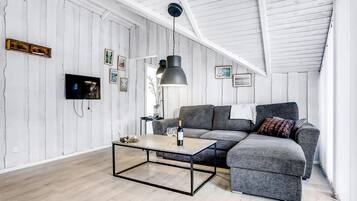 Un loft moderno con vigas de madera a la vista, un sofá seccional gris, una mesa de centro de vidrio y un televisor de pantalla plana montado en la pared.