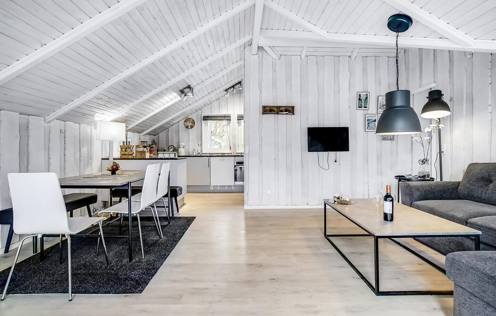 Amplia sala de estar con mesa de comedor, sillas, sofá y una mesa de centro. Se observa una cocina al fondo con un televisor montado en la pared.