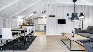 Un vaste espace de vie comprenant une table à manger, des chaises, un canapé et une table basse. On aperçoit une cuisine à l’arrière-plan, avec un téléviseur fixé au mur.