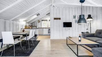 Amplia sala de estar con mesa de comedor, sillas, sofá y una mesa de centro. Se observa una cocina al fondo con un televisor montado en la pared.
