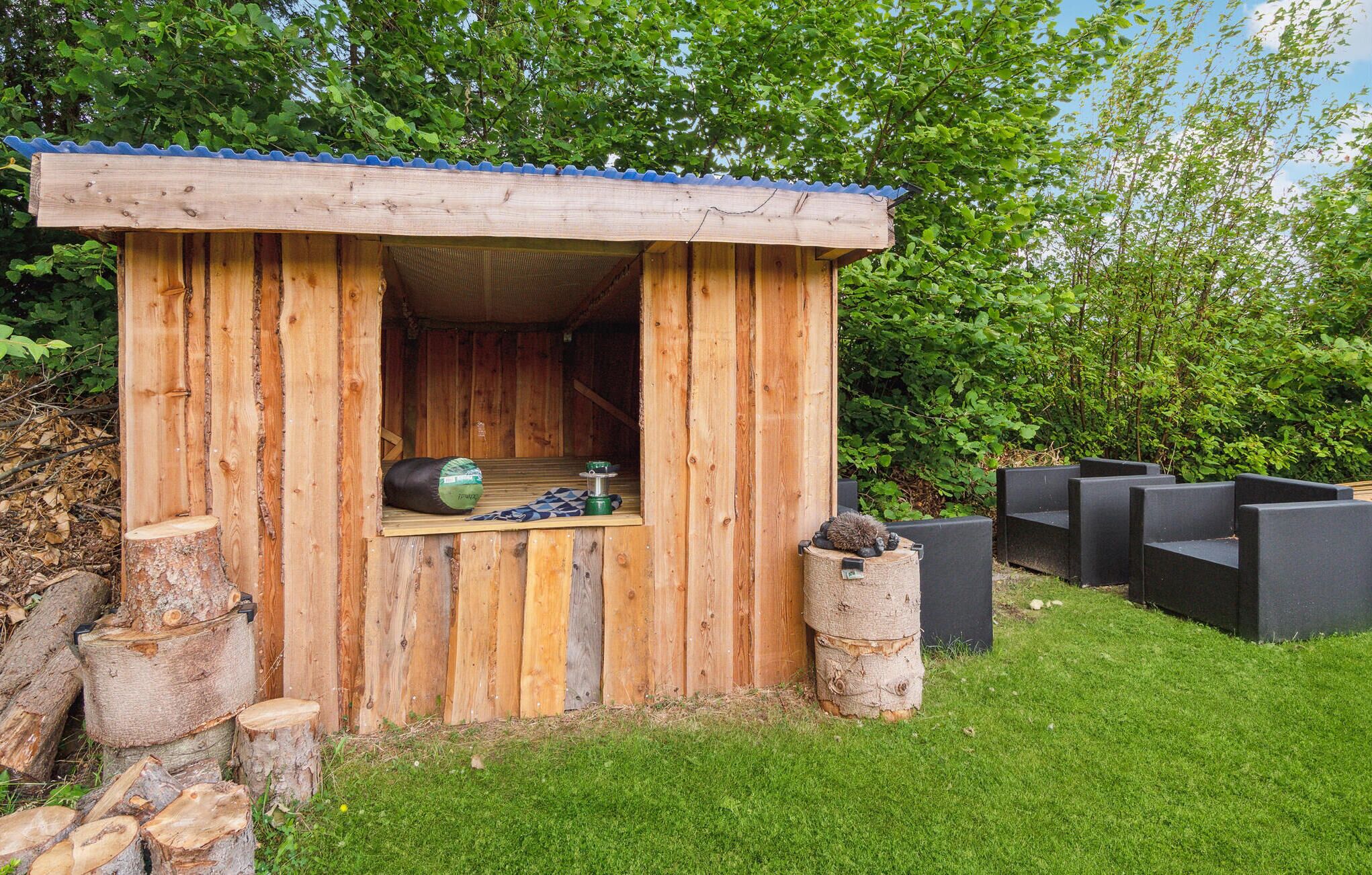 A wooden cabin with a blue roof, a small window, and a door. There are logs and a trash bin outside.