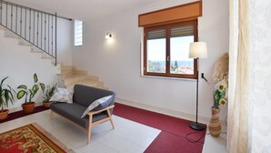 En stue med sofa, trappe, et vindue med rullegardiner, lampe og en vase med blomster.