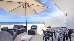 En udendørs terrasse med et spisebord, omgivet af fletmøbler, og en parasol der giver skygge.
