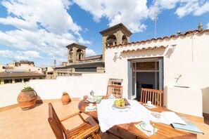 A rooftop terrace with a dining table set for two, surrounded by terracotta pots and a view of a historic building in the background.