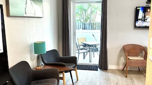 Quarto com piso de madeira, televisão fixada na parede, mesa de jantar com cadeiras e uma cadeira de vime.